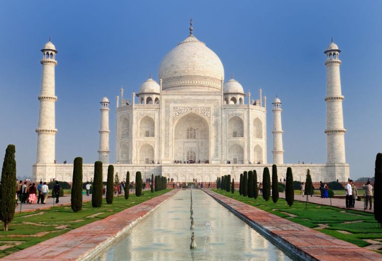 De witte marmeren Taj Mahal weerspiegeld in de langgerekte vijver, omringd door tuinen en bezoekers onder een heldere blauwe hemel.